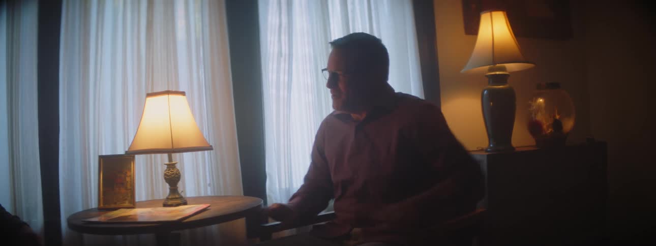 Two men are sitting while having a conversation stand up shake hands and embrace each other in a living room on an overcast day