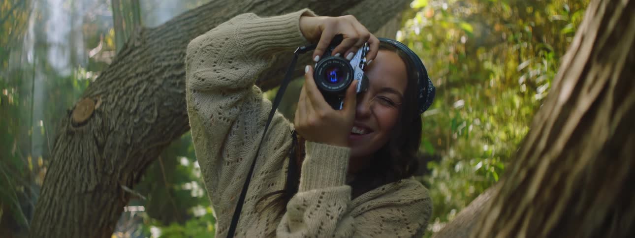 A woman smiling holding up a camera to take a picture in between trees in a forest