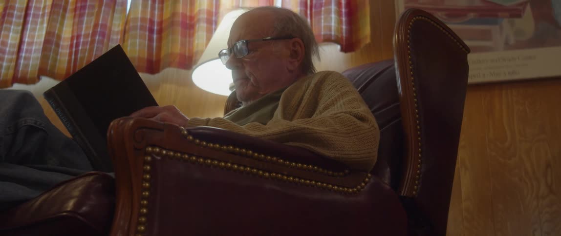 An elderly man reads a book while sitting in an armchair indoors during the day