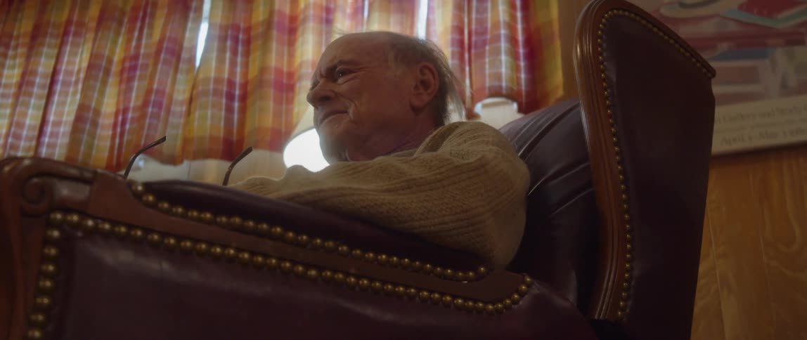 An elderly man is sitting in a chair as he holds his glasses in his hand and closes the book in his lap as he appears to be upset