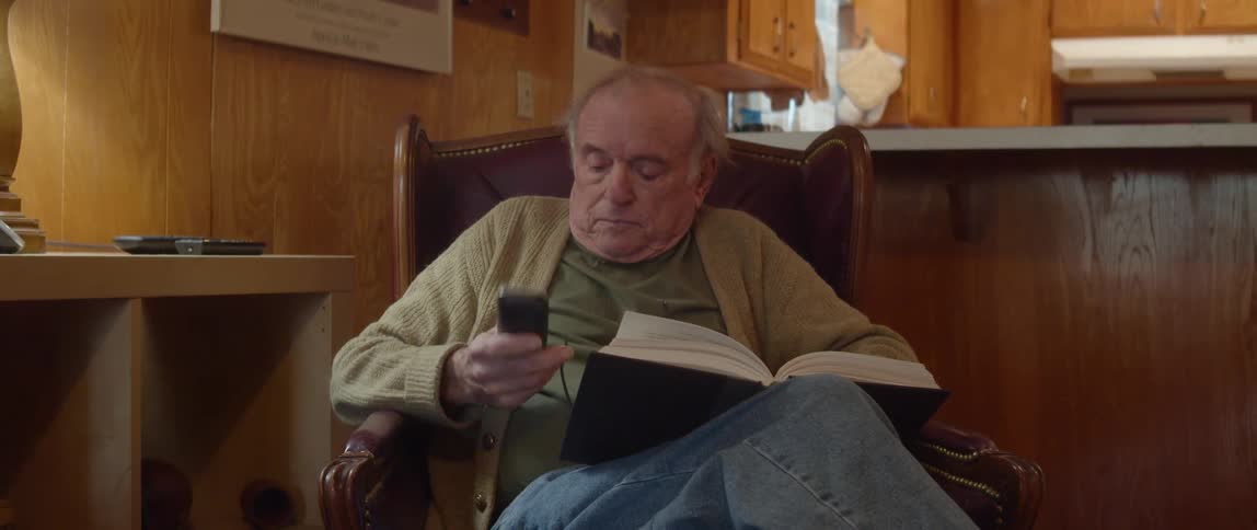 Elderly man with jeans green shirt and brown cardigan answers phone while reading a book in brown armchair