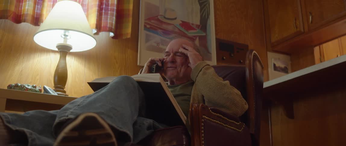 An elderly man is sitting in a chair in his living room with an open book in his lap smiling and laughing as he talks on the phone