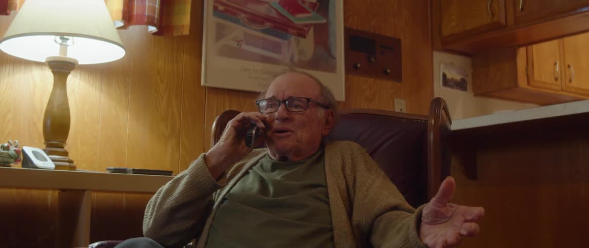 An elderly man is sitting in a chair in his living room smiling and laughing as and stretching his hand out as he talks on the phone