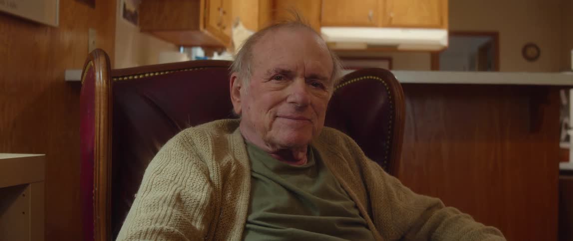 Elderly man with cardigan and shirt reads book and looks at camera in room