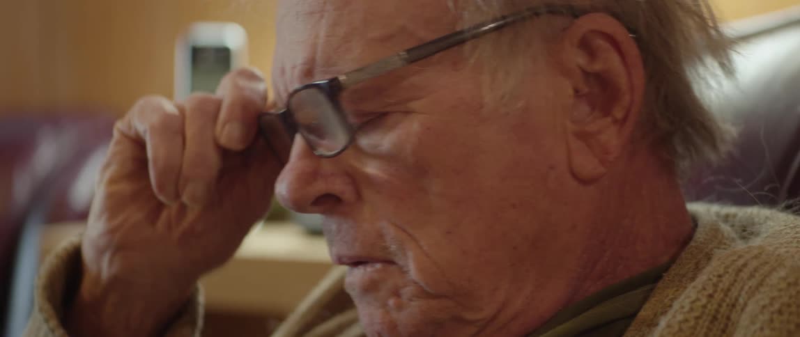 An elderly man is sitting in a chair in his living room as he puts on his glasses