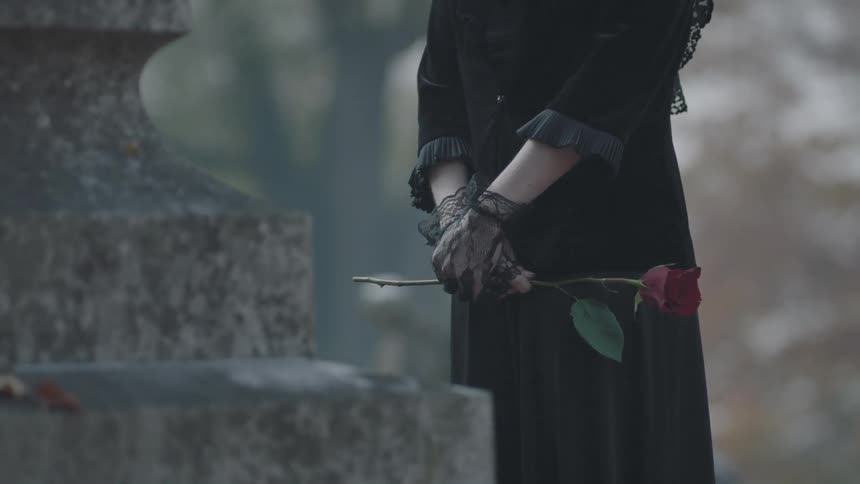 A woman holding a rose stands in front of a headstone