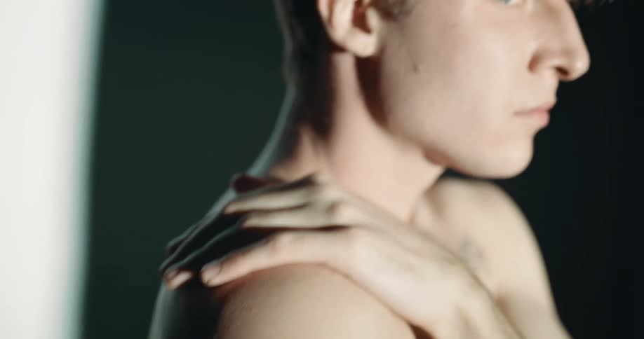 Young nude effeminate white man walks in and out of shadow and light while touching his right shoulder with his left hand