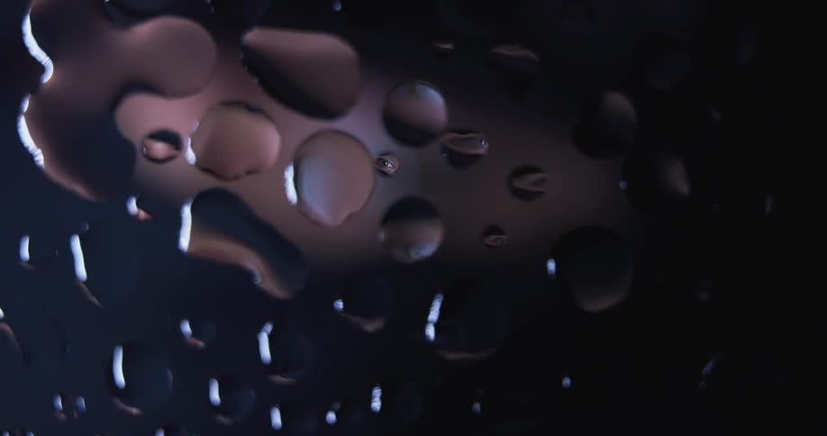 Close up of water or clear resin droplets on glass reflecting bits of a blinking eye thats behind the glass light source is camera left
