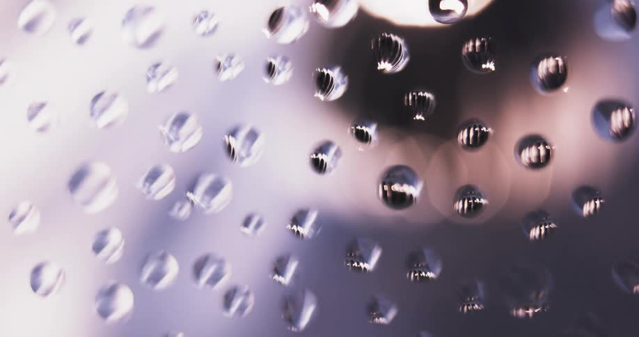 Water droplets sit on clear glass as pink light moves in the distance reflecting on the drops