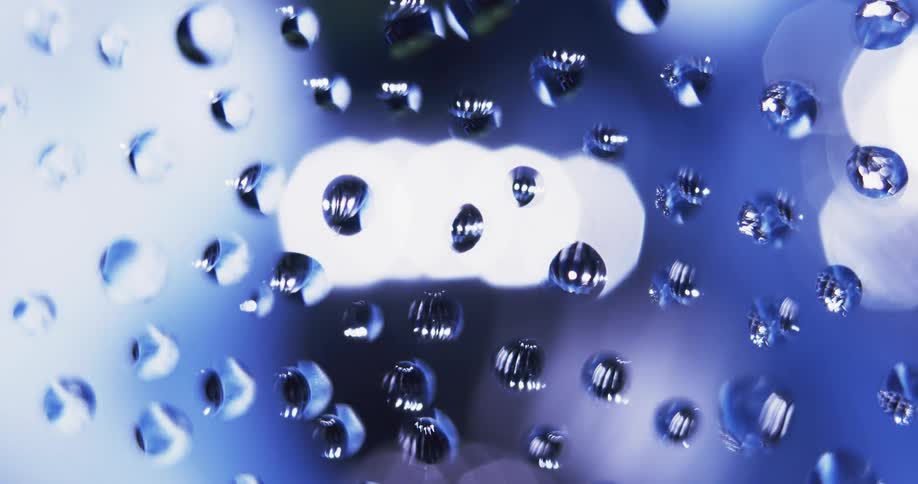 Water droplets sit on clear glass as blue and white light moves in the distance reflecting on the drops