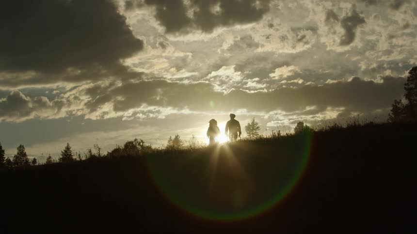 Man and woman walking toward horizon are silhouetted against sun under a cloudy sky