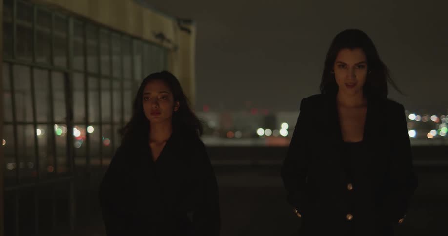 Two women in black walk past a building with many windows and there is a city lit up behind them at night