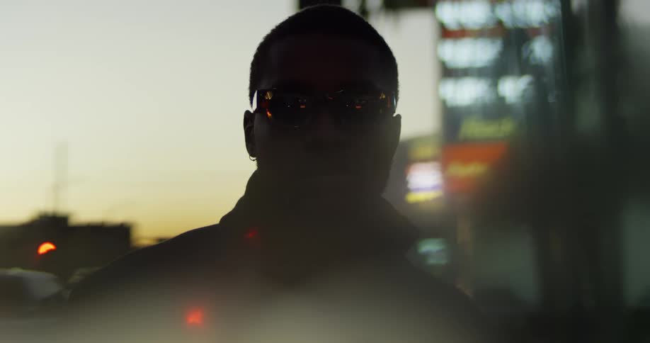 A man stands outside a city at dusk wearing sunglasses