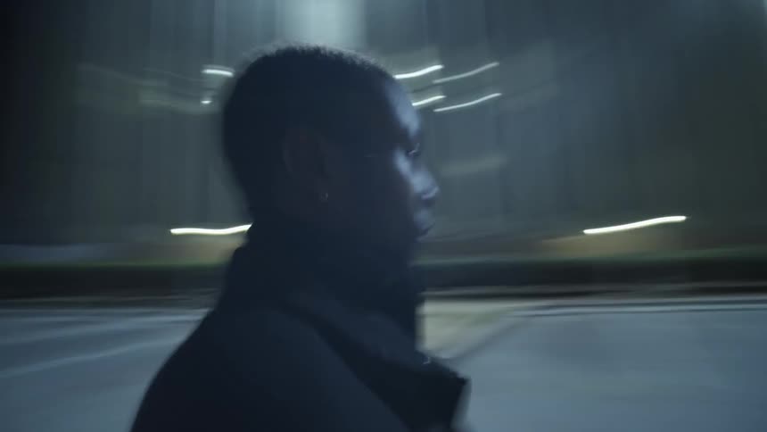 Man with black overcoat and earrings stands still and stares into space next to tall building at night in the city