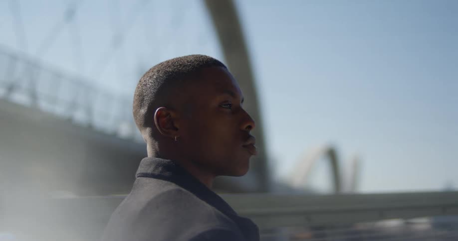 Man with earrings black overcoat looks around near bridge with city skyline in the background