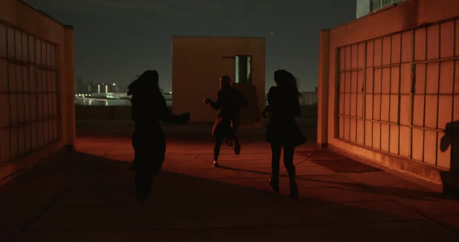 Three young people a man and two women are having fun running on the rooftop of a building at night laughing while the city skyline and lights can be seen in the background