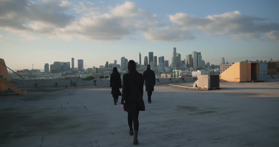 Man in black coat and women wearing black coat and blazer jacket walk on top of the building with city skyline in the background