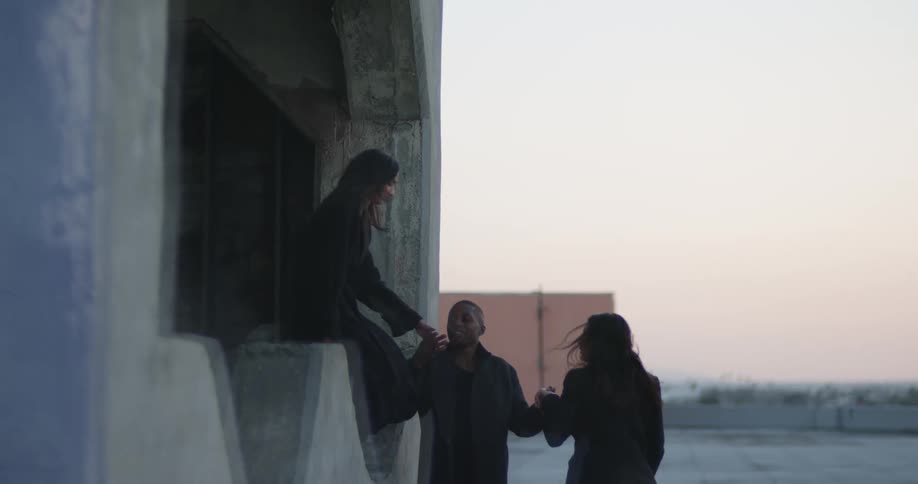 A woman walks past a window to ledge of a roof building where as man is standing as he helps her jump down another woman appears and grabs his other hand as she steps of the ledge and then they all run towards the roof access door as the sun is setting