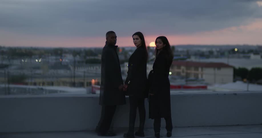 Three people two white women one black man all in black trench coats standing on the roof of a building at sunset in big city looking over shouler at camera