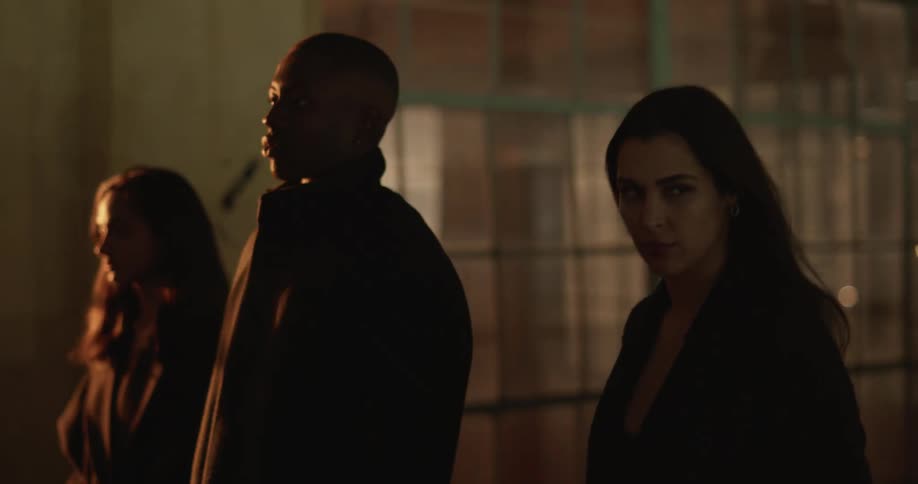 Three people strut past a building as something explodes in the distance