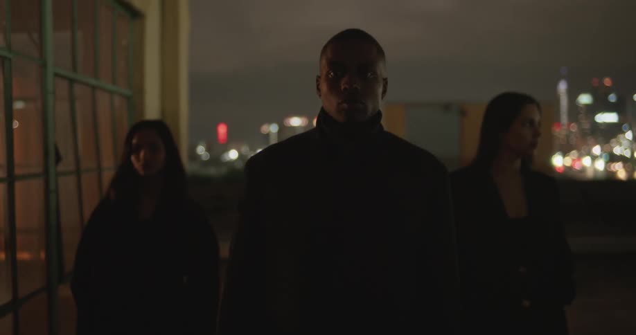 Man in black coat and women wearing black coat and blazer jacket walk on top of building with city skyline in the background at night