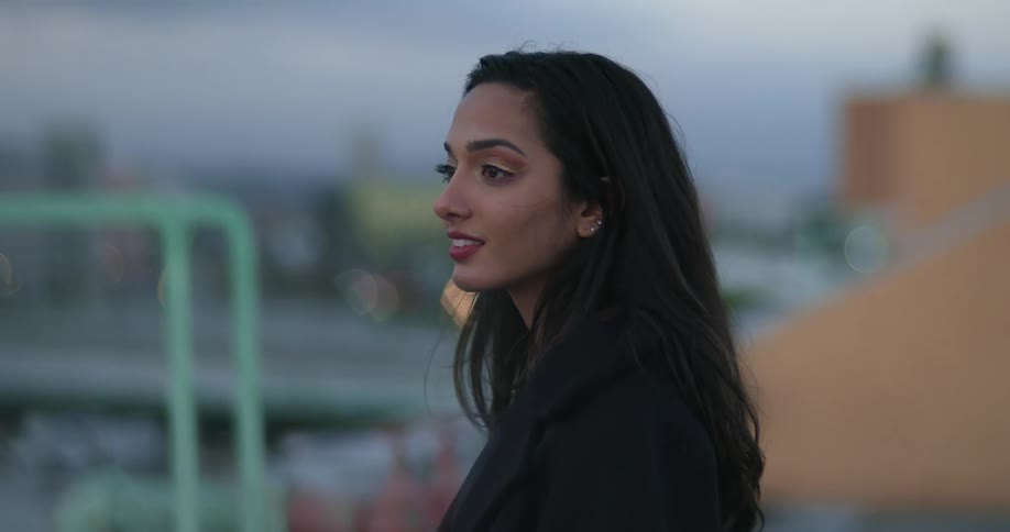 Woman wearing fashionable clothes stands on building roof smiling slightly as she looks across roof toward another person