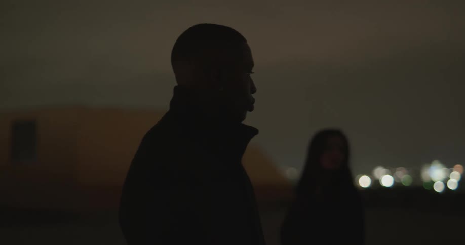 At nighttime a man and two women in black walk in circles around one another with a city lit up in the background
