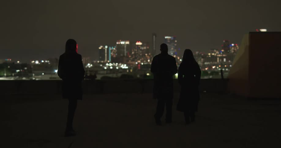 Three people walk across roof with view of skyline at night as spotlight move across area