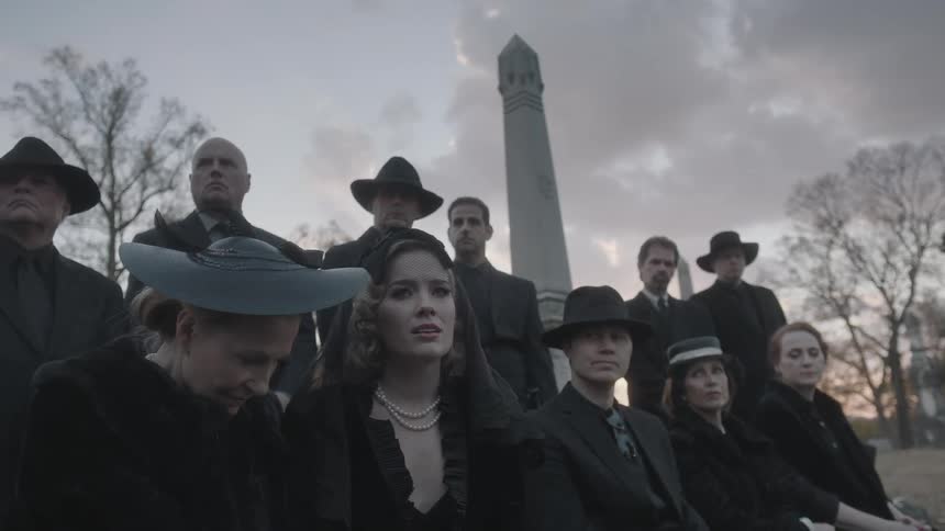 A group of mourners in dark clothing sit in a cemetery