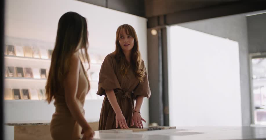 Two women are standing in a shop over a table as they talk and point to items on the table