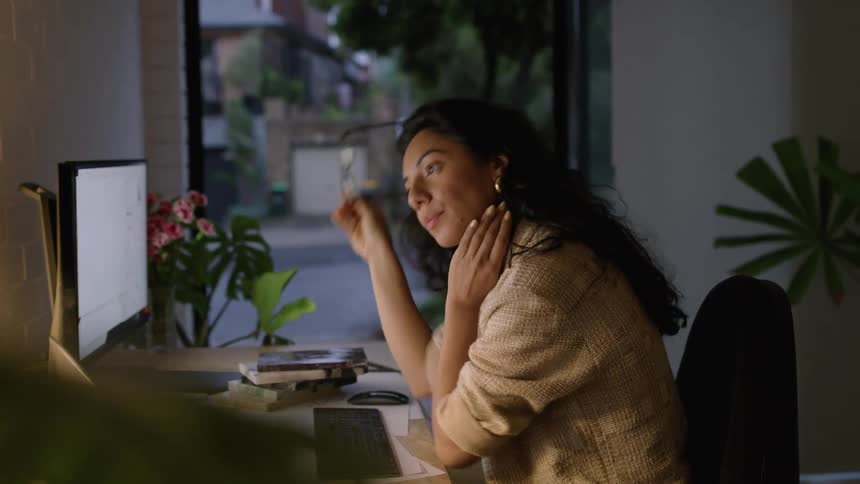 A woman is sitting indoors at a desk doing work at a computer at dusk