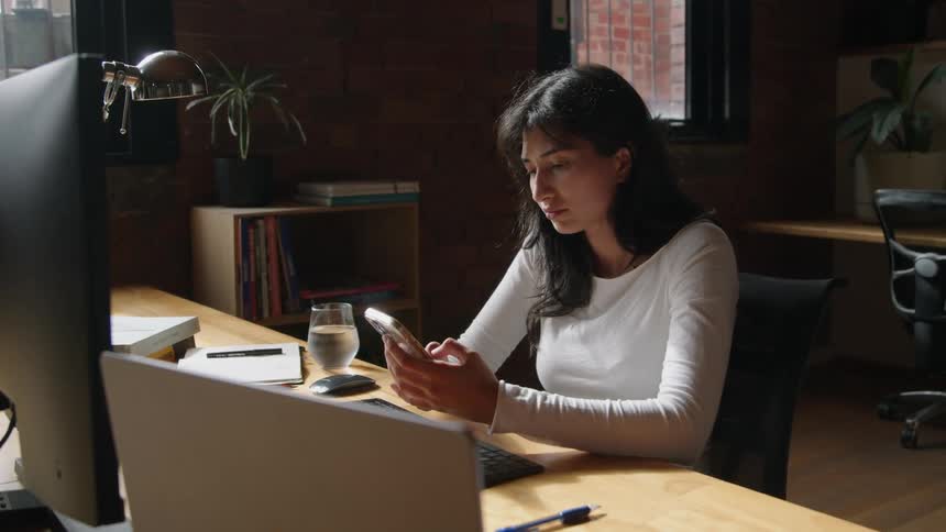 A woman using a computer at a desk stops to check her phone
