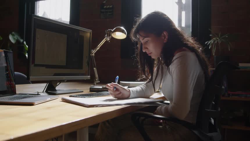 A woman is writing on a notebook and sitting at her desk