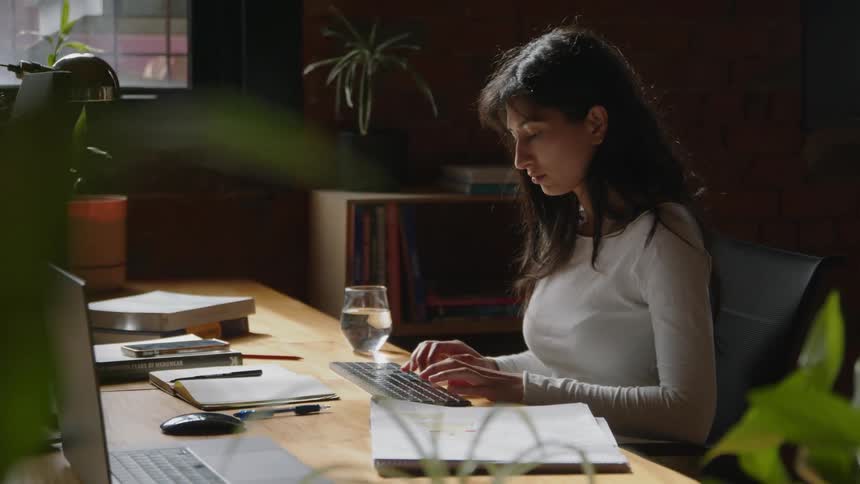 Woman with dark hair and white shirt sits at desk tying on computer keyboard in the office