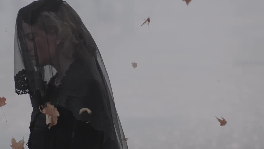 A woman wearing a lace hat mourns as leaves fly around her