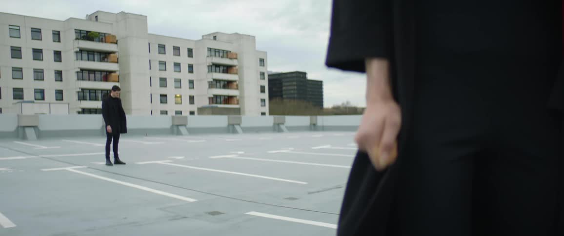 A woman wearing black clothes is standing in a parking lot with a clenched fist as a man wearing black is standing behind her holding his phone and there are buildings in the distance