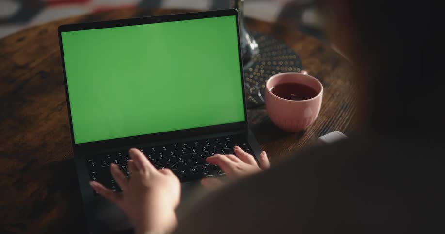A cup of coffee sits next to a laptop computer with a green screen on a table as a woman begins to type on the keyboard