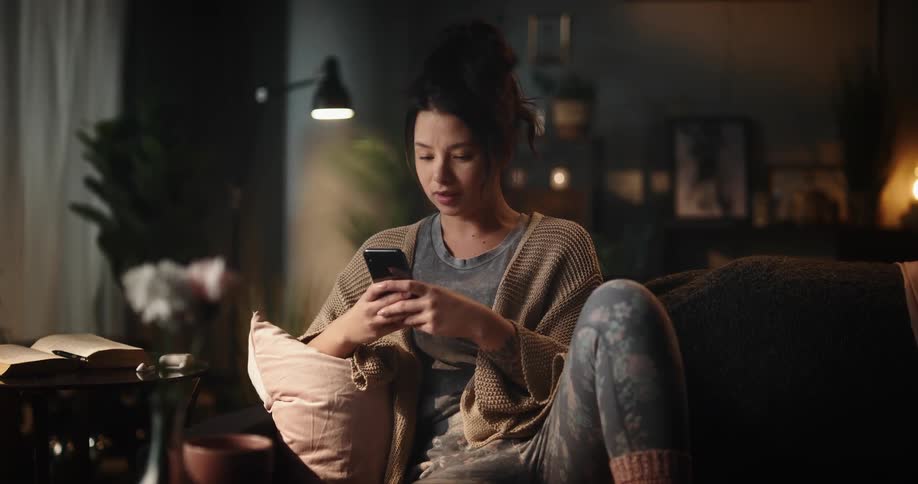 Woman shakes her head and frowns while sitting on couch and looking at her smartphone screen