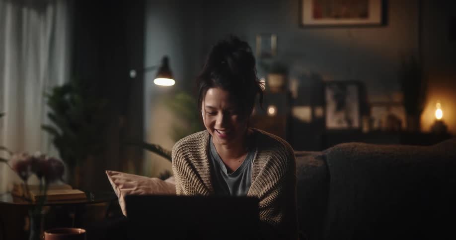 Woman sits on couch in dimly lit living room holding conversation via laptop