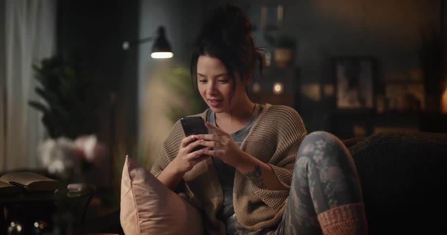 A woman laughs while scrolling through her phone on the couch