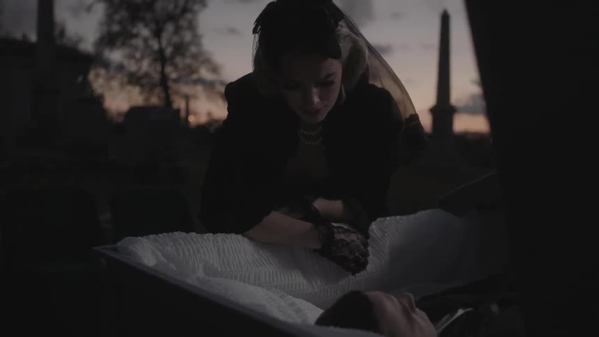 A woman wearing a lace hat mourns looking down at a deceased man in a casket