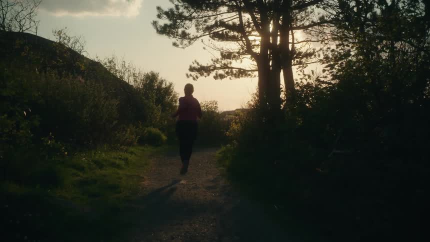 Woman with pink jacket and black leggings jogs along track in the forest at sunset