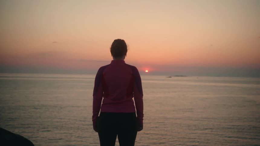 Woman with pink jacket and black leggings runs to the edge of a cliff overlooking ocean and sunset