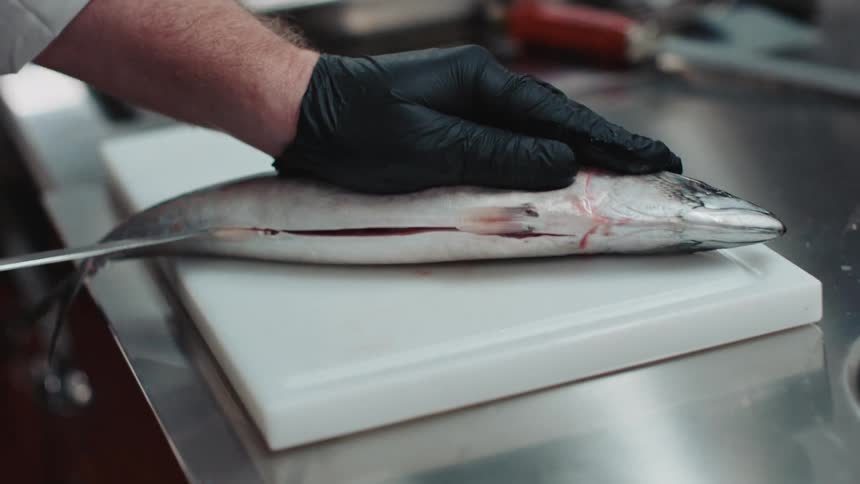 Chef wearing black latex gloves uses a sharp kitchen knife to slice and cut a fish