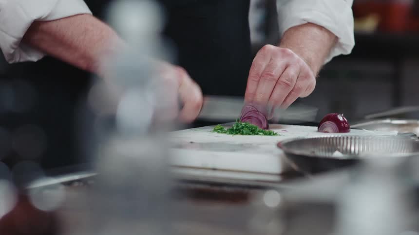 Cook with white shirt and black apron cuts onion on cutting board with knife