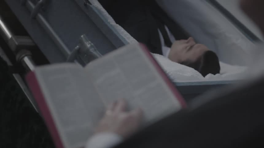 A man reads from a bible as a deceased man lays in a casket