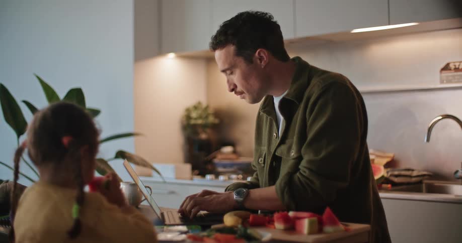 A man types on his laptop in the kitchen as his daughter eats watermelon sitting next to him