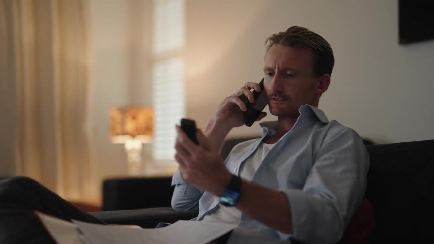 A man in a room on his bed making a call reading through a book and switching off the tv using a remote