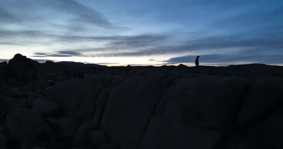 Aerial view of man with cap and jacket walking on rocky mountain is being silhouetted against the sky at dusk
