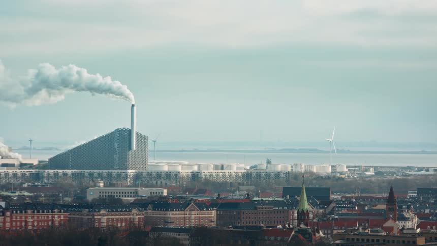 Smoke rises from chimney at manufacturing plant towering over nearby buildings with foggy water in background